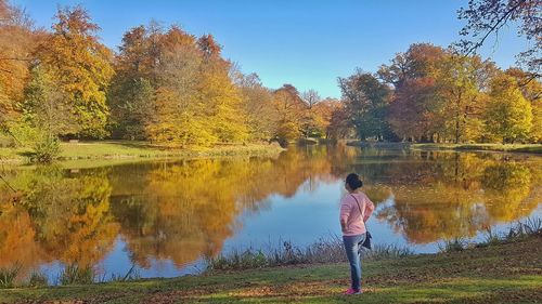 Rear view of man standing by lake against sky during autumn