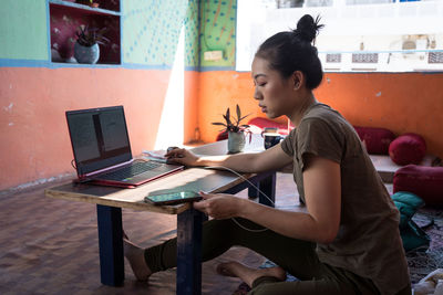 Side view of busy female freelancer sitting on floor at table with laptop in shabby room and browsing cellphone while working remotely on project