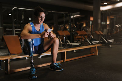 Full length of young man with prosthetic leg using phone while sitting on bench in gym