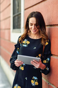 Smiling young woman using digital tablet while standing by wall