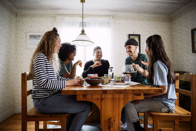 Happy friends talking while enjoying food and drink on table at home