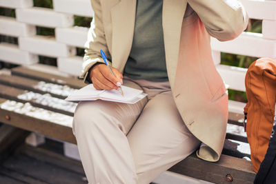 Cropped unrecognizable entrepreneur in suit sitting on bench and writing in planner while working remotely on sunny day in street restaurant