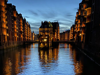 Canal amidst illuminated buildings against sky at sunset