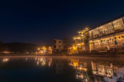 Illuminated buildings by lake against sky in city at night