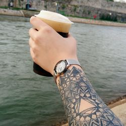 Cropped hand with mandala tattoo holding beer glass over river
