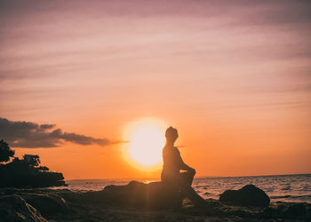 Side view of man on rock at beach against sky during sunset