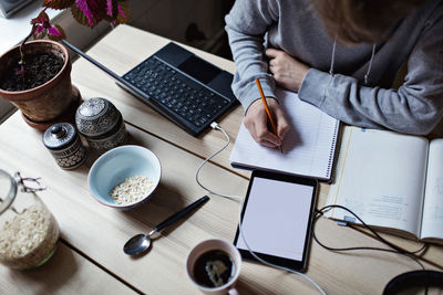 High angle view of teenage boy writing on book while using digital tablet for homework