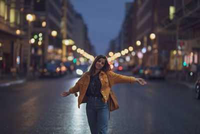 Portrait of young woman standing with arms outstretched on road in city at night