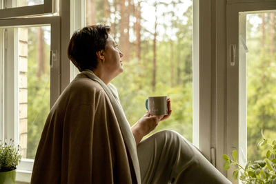 Middle age beautiful woman sitting on windowsill, drinking tea, dreaming. 50-year-old woman relaxing