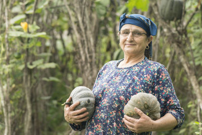 Portrait of smiling senior woman holding pumpkins against trees