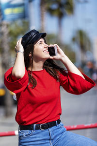 Smiling teenage girl wearing red top while using mobile phone in city during sunny day