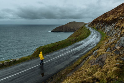 High angle view of man wearing yellow raincoat walking on road by sea against cloudy sky