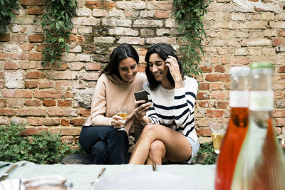 Smiling female friends sharing smart phone sitting on bench against brick wall at garden party