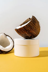 Two halves of coconut and a jar of cosmetics on a yellow and gray background.