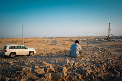 Rear view of man sitting on sand at beach against clear sky