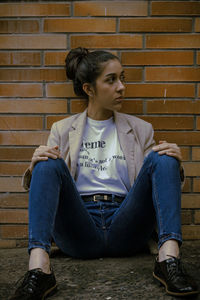 Young woman looking away while sitting against brick wall