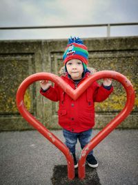 Portrait of boy standing behind red heart shape against railing