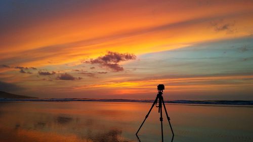 Silhouette tripod on shore at beach against sky during sunset
