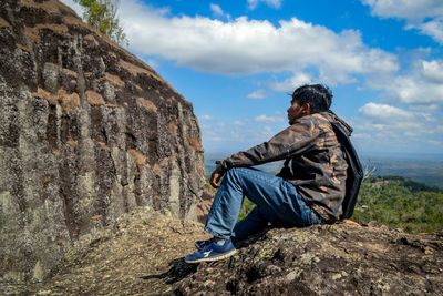 Side view of man sitting on rock against sky