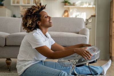 Happy young black woman sits in front of fan at home cooling down at home after being in heat