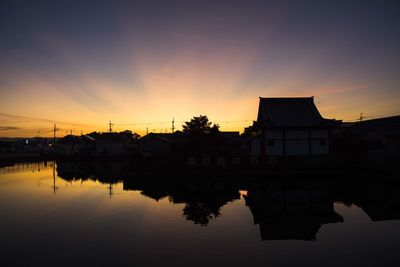 Silhouette buildings by lake against sky during sunset