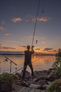 Rear view of man fishing in sea against sunset sky