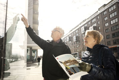 Senior woman reading catalog while man pointing on glass window in city