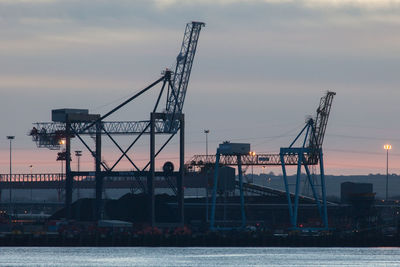 Cranes at commercial dock against cloudy sky at dusk