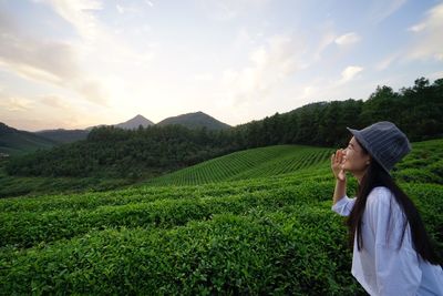 Side view of young woman standing on tea field against sky during sunset