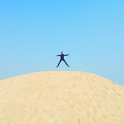 Low angle view of man with arms outstretched jumping over sand at beach against clear blue sky