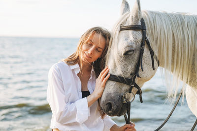 Young long hair woman in white shirt with white horse on seascape background