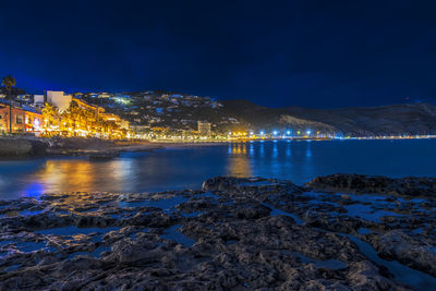 Illuminated buildings by sea against sky at night