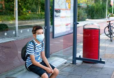 A schoolboy sitting waiting for a bus wearing protective face mask during quarantine, back to school 