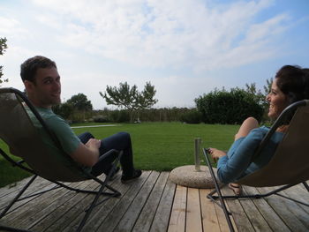 Man with female friend sitting on chairs by grassy field against sky