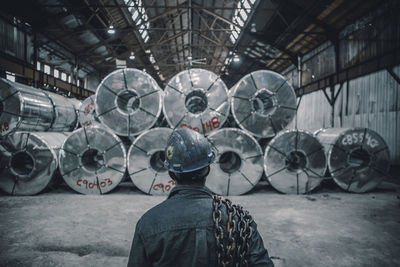 Rear view of male worker carrying chain standing in front of rolled up steel sheets at factory