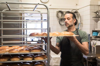 Confident mature male owner holding baking sheet with freshly baked breads on rack at bakery kitchen