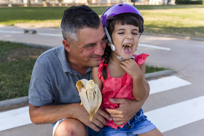 Father and daughter eating banana sitting outdoors on the grass in a park.