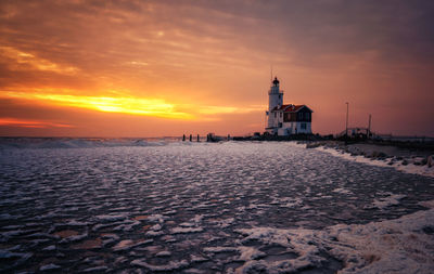 Lighthouse amidst frozen sea against cloudy sky during sunset