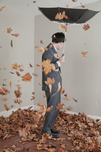 Man holding damaged umbrella while standing by dry leaves against wall