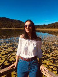 Portrait of young woman wearing sunglasses while standing by lake against blue sky