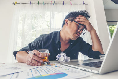 Depressed businessman holding whiskey glass and cigarette while sitting with head in hands at office desk