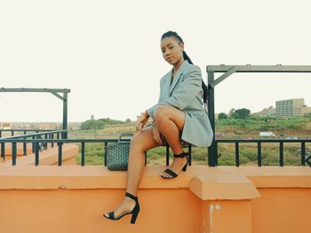 Young woman sitting on railing in city against clear sky
