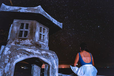 Woman sitting by built structure against sky at night