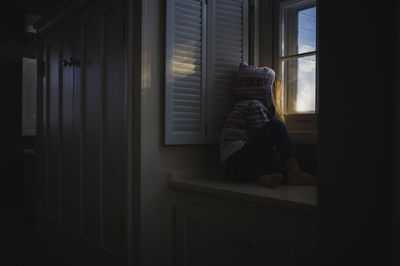 Girl in hooded shirt looking through window while sitting on cabinet at home