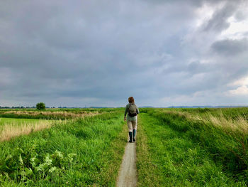 Rear view of woman walking over a rural footpath between meadows under grey sky