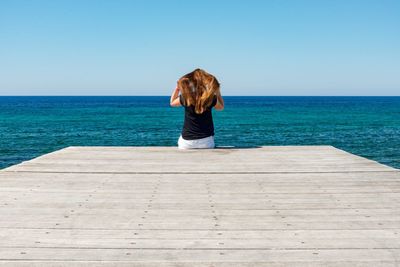 Rear view of woman with hands in hair sitting on jetty over sea against clear sky