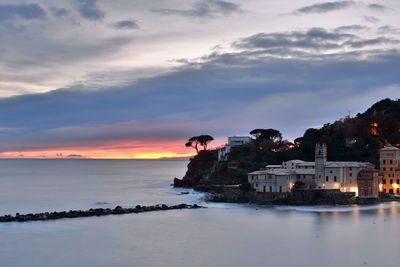 Sea by illuminated buildings against sky at sunset