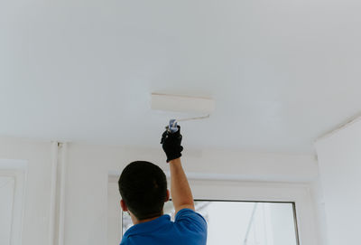 A young oriental man paints the ceiling near the window with a roller.