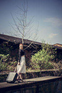 Woman standing while holding luggage against sky