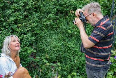 Midsection of man photographing woman standing by plants